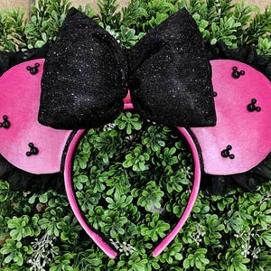 May include: A pink headband with large pink circular ears adorned with black Mickey Mouse-shaped embellishments. A large, sparkly black bow sits atop the ears. Black lace trim surrounds the ears.
