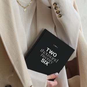 May include: A black notebook with the text "TWO thousand twenty SIX" in white is held against a beige blazer. The blazer has brown buttons and is worn over a cream-colored sweater. A gold necklace with a pendant is visible.