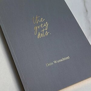 May include: A gray notebook with the text "the grey dots." in gold lettering. The text "Dein Wunschtext" is also in gold lettering.