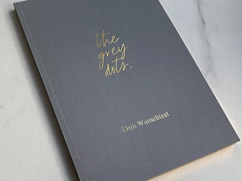 May include: A gray notebook with the text "the grey dots." in gold lettering. The text "Dein Wunschtext" is also in gold lettering.
