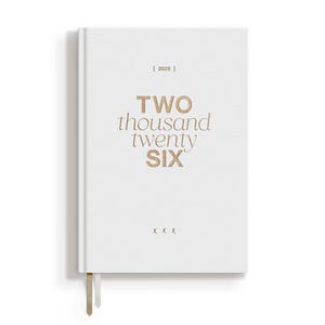 May include: A white hardcover journal with the text "TWO thousand twenty SIX" in gold lettering. The year "2026" is printed above the title. A gold ribbon bookmark hangs from the bottom edge.