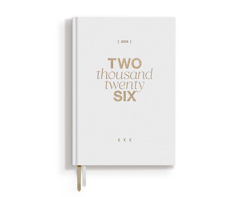 May include: A white hardcover journal with the text "TWO thousand twenty SIX" in gold lettering. The year "2026" is printed above the title. A gold ribbon bookmark hangs from the bottom edge.