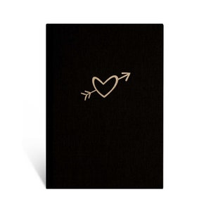 May include: A black notebook with a gold heart pierced by an arrow design. The heart and arrow are centered on the front cover. The notebook is standing upright against a white background.