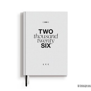 May include: A white hardcover journal with the text "TWO thousand twenty SIX" in black. The journal has a black ribbon bookmark and the year "2026" printed at the top. Three small "x" marks are at the bottom.
