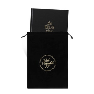May include: A black velvet pouch with a gold drawstring closure. The pouch has a gold circle logo that reads "Luxury Paper & Design Goods Real Passionates www.realpassionates.com". The pouch contains a black leather-bound journal with gold lettering that reads "the killer ideas".