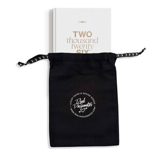 May include: A white 2026 planner with gold lettering reading "TWO thousand twenty SIX" is partially visible behind a black drawstring bag. The bag has a white logo that reads "Real Passionates" and "Luxury Paper & Design Goods."