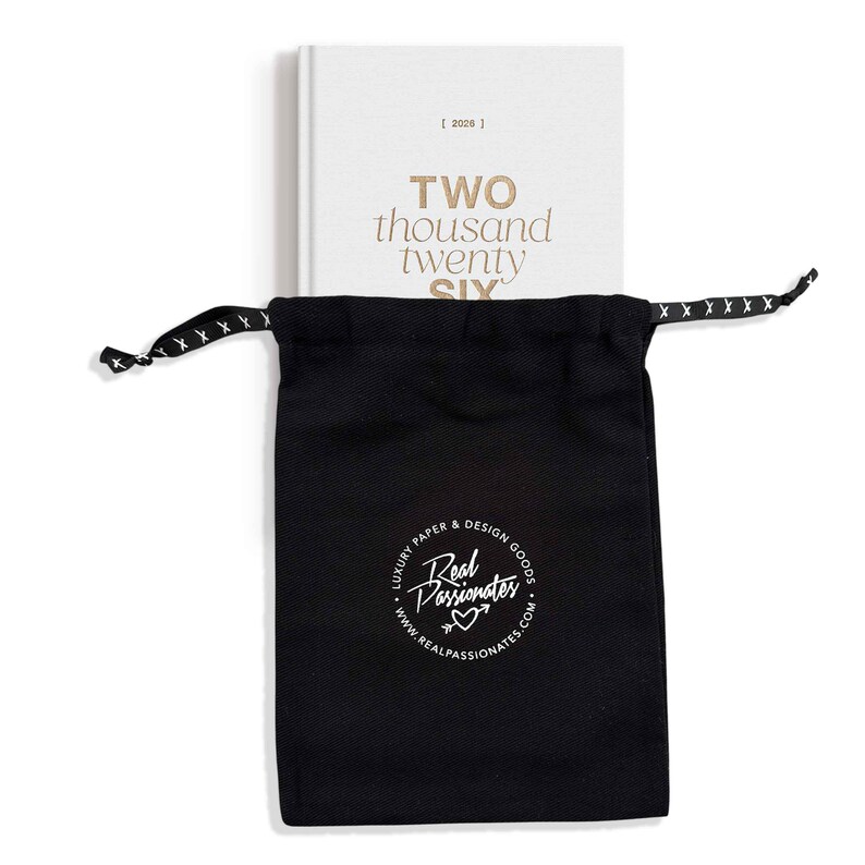 May include: A white 2026 planner with gold lettering reading "TWO thousand twenty SIX" is partially visible behind a black drawstring bag. The bag has a white logo that reads "Real Passionates" and "Luxury Paper & Design Goods."