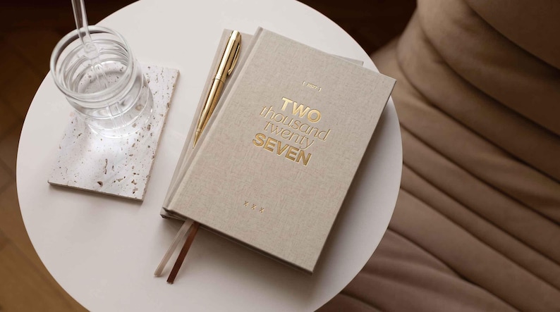 May include: A beige fabric-covered planner with the gold embossed text TWO THOUSAND TWENTY SEVEN on the front cover, accompanied by a gold metal pen and two thin ribbon bookmarks, resting on a white circular table next to a glass of water.