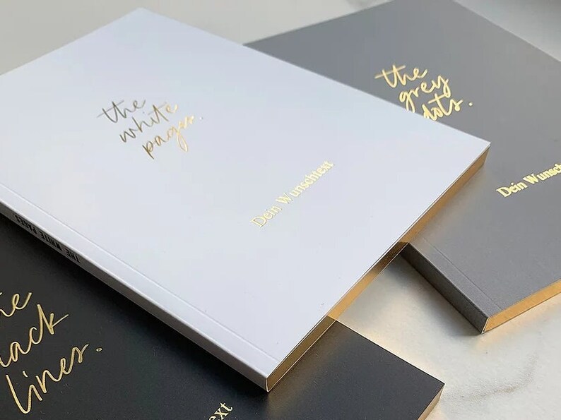 May include: Three notebooks with gold foil lettering. The white notebook says "the white pages" and "Dein Wunsch", the gray notebook says "the grey dots" and "Dein Wunsch", and the black notebook says "the black liner" and "Dein Wunsch".
