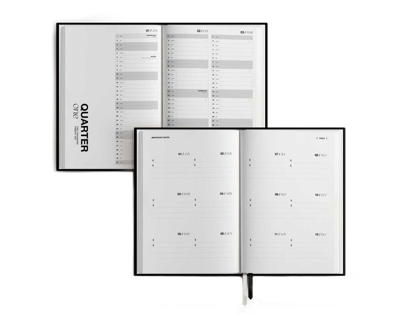 May include: Two open black-bound planners with white pages. The top planner displays a yearly calendar layout, while the bottom planner shows a monthly format. The text "Quarter One" is printed on the side of the top planner.