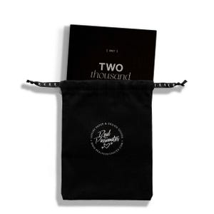 May include: A black drawstring pouch featuring a white circular logo with the text Real Passionates and a heart arrow icon. Inside the pouch is a black book with the text 2027 and TWO thousand printed in white on the front cover.