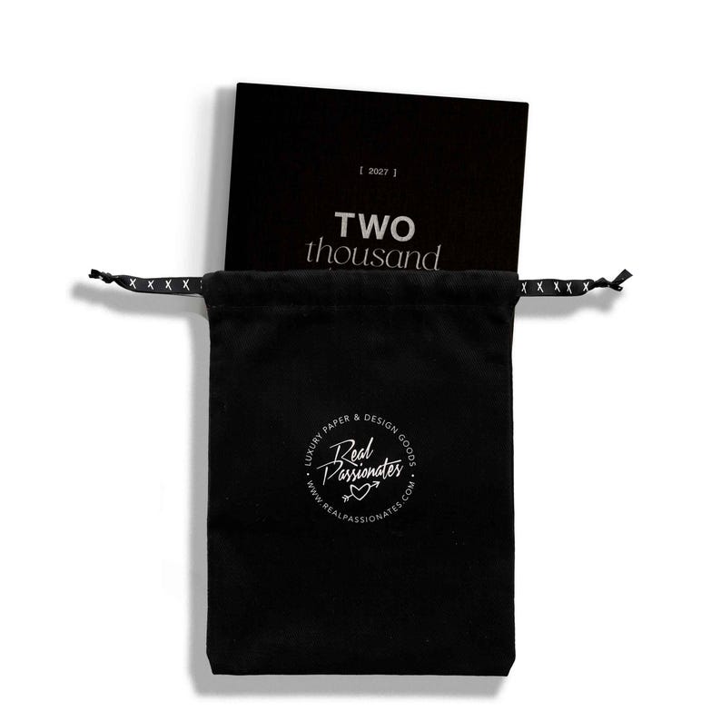 May include: A black drawstring pouch featuring a white circular logo with the text Real Passionates and a heart arrow icon. Inside the pouch is a black book with the text 2027 and TWO thousand printed in white on the front cover.