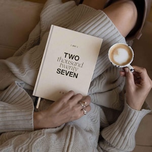 May include: A cream-colored hardcover planner with the text TWO thousand twenty SEVEN printed in black on the front cover. The planner is held by a person wearing a beige ribbed sweater, who is also holding a small cup of coffee with latte art.