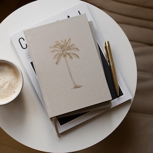 May include: A beige journal with a gold palm tree illustration on the cover. The journal is stacked on top of a magazine with the letter "C" visible. A gold pen is lying on top of the journal and magazine. A cup of coffee is in the foreground.