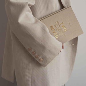 May include: A beige blazer with four buttons on the sleeve cuff, paired with a beige hardcover book featuring the gold embossed text TWO THOUSAND TWENTY SEVEN on the front cover. The book has a textured fabric finish and a clean, neutral appearance.