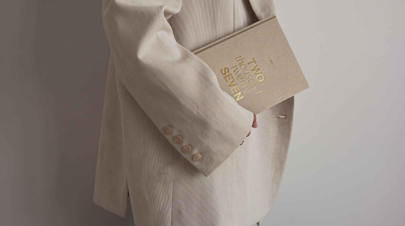 May include: A beige blazer with four buttons on the sleeve cuff, paired with a beige hardcover book featuring the gold embossed text TWO THOUSAND TWENTY SEVEN on the front cover. The book has a textured fabric finish and a clean, neutral appearance.