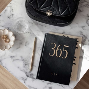 May include: A black leather journal with gold lettering that says "365" on the cover. The journal is sitting on a white marble surface with a gold pen, a glass cup, and a small white dish with gold rings.