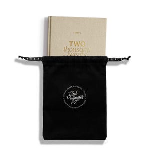 May include: A beige hardcover planner labeled 2027 and TWO thousand twenty, partially inside a black drawstring pouch. The pouch features a white circular logo with the text Real Passionates, LUXURY PAPER & DESIGN GOODS, and a website URL.