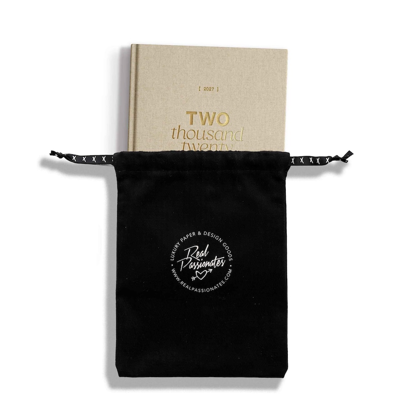 May include: A beige hardcover planner labeled 2027 and TWO thousand twenty, partially inside a black drawstring pouch. The pouch features a white circular logo with the text Real Passionates, LUXURY PAPER & DESIGN GOODS, and a website URL.