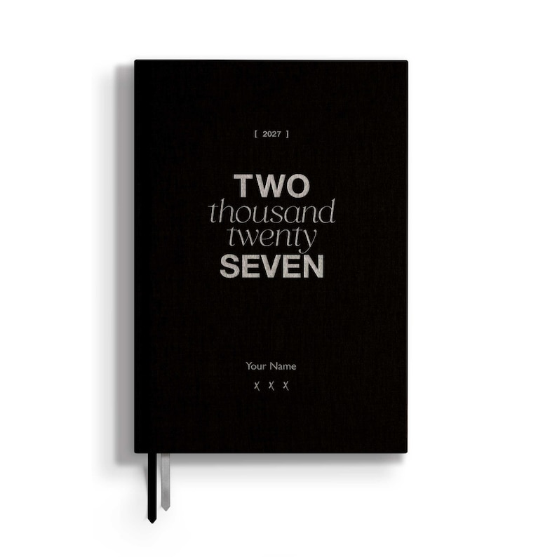 May include: A black rectangular planner with a textured cover featuring the text [ 2027 ] at the top, the words TWO thousand twenty SEVEN in the center, and the placeholder text Your Name followed by three X characters at the bottom.