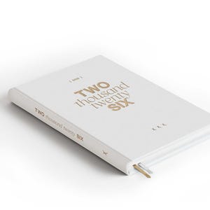 May include: A white hardcover journal with the text "TWO thousand twenty SIX" in gold lettering. The journal has a gold and silver ribbon bookmark. The cover also has the text "TWO thousand twenty SIX" on the spine.