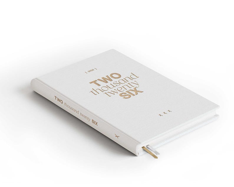 May include: A white hardcover journal with the text "TWO thousand twenty SIX" in gold lettering. The journal has a gold and silver ribbon bookmark. The cover also has the text "TWO thousand twenty SIX" on the spine.