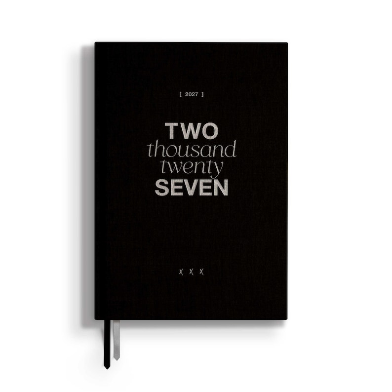 May include: A black planner with the text 2027 in brackets at the top, the words TWO thousand twenty SEVEN in the center, and three X symbols at the bottom. The cover has a textured finish and two thin ribbon bookmarks extending from the bottom edge.