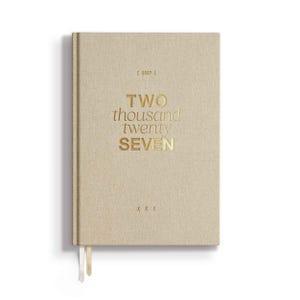 May include: A beige fabric-covered planner with gold foil lettering on the front cover that reads 2027, TWO thousand twenty SEVEN, and X X X. The planner features two thin ribbon bookmarks extending from the bottom edge of the hardbound cover.