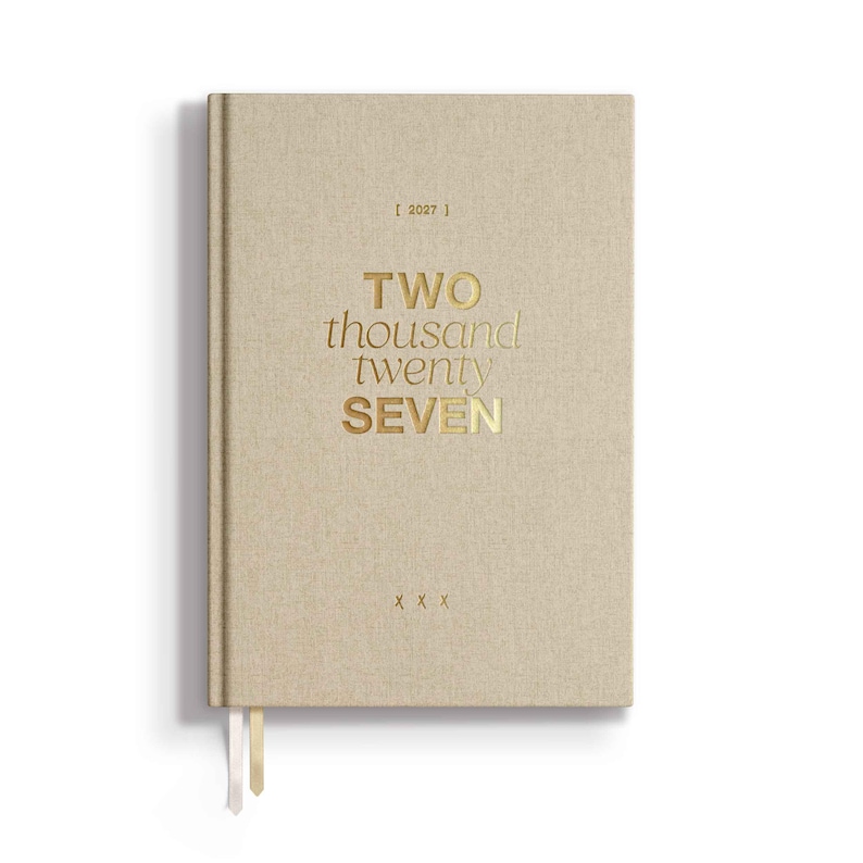 May include: A beige fabric-covered planner with gold foil lettering on the front cover that reads 2027, TWO thousand twenty SEVEN, and X X X. The planner features two thin ribbon bookmarks extending from the bottom edge of the hardbound cover.
