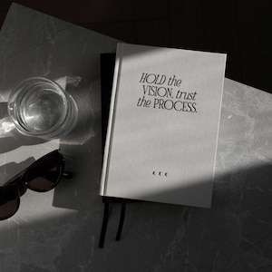 May include: A white notebook with the text "HOLD the VISION, trust the PROCESS." printed in black. The notebook is lying on a gray surface with a glass of water and sunglasses.