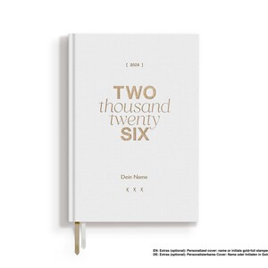 May include: A white hardcover journal with the text "TWO thousand twenty SIX" in gold lettering. The journal includes a bookmark and space for a name. The year "2026" is printed at the top.