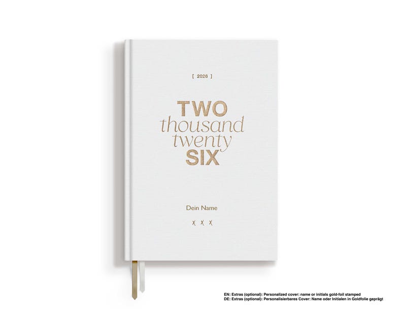 May include: A white hardcover journal with the text "TWO thousand twenty SIX" in gold lettering. The journal includes a bookmark and space for a name. The year "2026" is printed at the top.