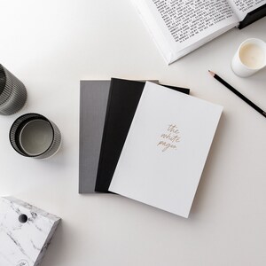 May include: A white notebook with the text "the white pages." written in gold lettering. The notebook is on a white surface with a black pencil, a glass candle holder, and a gray and white marble box.