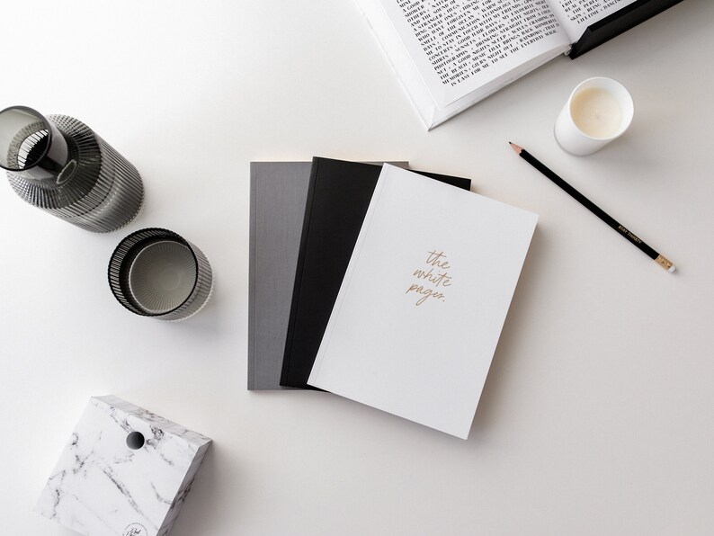 May include: A white notebook with the text "the white pages." written in gold lettering. The notebook is on a white surface with a black pencil, a glass candle holder, and a gray and white marble box.