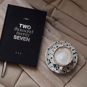 May include: A black hardcover planner with the silver text TWO thousand twenty SEVEN on the cover, accompanied by a white ceramic cup and saucer featuring a black speckled pattern, both resting on a rectangular light beige stone board.