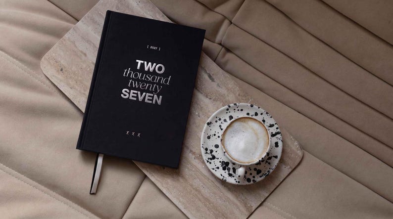 May include: A black hardcover planner with the silver text TWO thousand twenty SEVEN on the cover, accompanied by a white ceramic cup and saucer featuring a black speckled pattern, both resting on a rectangular light beige stone board.