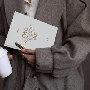 May include: A person holding a white notebook with the text "TWO thousand twenty SIX" in gold lettering, a gold pen, and a white coffee cup. The person is wearing a gray coat.