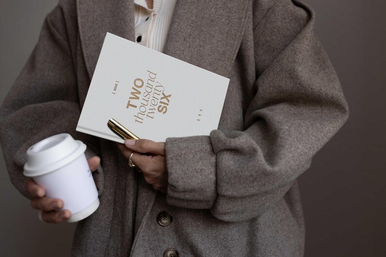 May include: A person holding a white notebook with the text "TWO thousand twenty SIX" in gold lettering, a gold pen, and a white coffee cup. The person is wearing a gray coat.