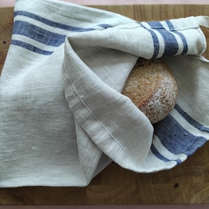 May include: A natural-coloured linen bread bag featuring two horizontal navy blue stripes. The fabric has a visible woven texture, and the bag is partially open, revealing a round, crusty loaf of brown bread tucked inside the pouch.