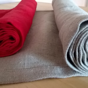May include: Two rolled fabric table runners, one red and one grey, are lying on a wooden surface. The red runner is rolled tightly, while the grey runner is rolled loosely.