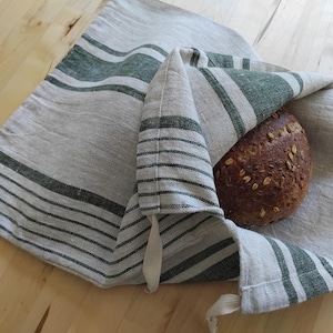 May include: A rectangular linen bread bag featuring a natural beige base with dark green horizontal stripes of varying widths. The bag is partially open, revealing a round, dark brown artisan loaf of bread topped with visible grains and seeds.