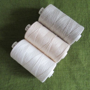 May include: Three spools of thread in different shades of beige, from light to dark, on a green fabric background.