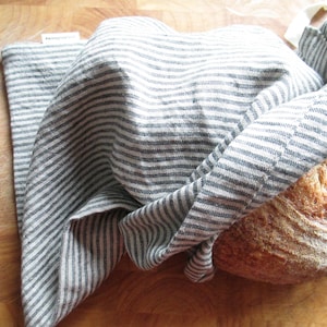 Op de afbeelding: Een versgebakken brood, gedeeltelijk verpakt in een grijs-wit gestreepte linnen broodzak. Het brood heeft een goudbruine korst. De zak is gemaakt van een zachte, getextureerde stof. De scène speelt zich af op een houten oppervlak.