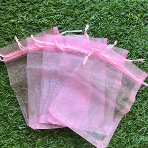 Puede incluir: Un conjunto de seis bolsas de regalo de organza rosa con lazos de cinta blanca. Las bolsas están apiladas una encima de la otra y están sobre una superficie de hierba verde.