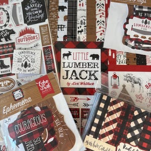 Op de afbeelding: Een verzameling scrapbookpapier en versieringen met een lumberjack-thema. De items bevatten papier met patronen, gestanste labels en stickers met beren, bijlen en buffelruitpatronen in rood, zwart en wit. Tekst bevat "Little Lumber Jack" en "Courageous."