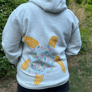 Puede incluir: Sudadera con capucha gris claro con un dise&ntilde;o de tortuga floral en la espalda. El caparaz&oacute;n de la tortuga es azul claro con flores rosas y amarillas, y las aletas son amarillas. La sudadera tiene capucha y es de material suave.