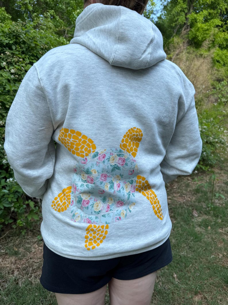 Puede incluir: Sudadera con capucha gris claro con un dise&ntilde;o de tortuga floral en la espalda. El caparaz&oacute;n de la tortuga es azul claro con flores rosas y amarillas, y las aletas son amarillas. La sudadera tiene capucha y es de material suave.