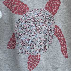 Puede incluir: Una sudadera gris con un dise&ntilde;o de tortuga marina. El caparaz&oacute;n de la tortuga tiene un estampado floral en rojo y blanco, mientras que las aletas y la cabeza son de color rojo liso. El dise&ntilde;o est&aacute; centrado en la parte delantera de la sudadera.