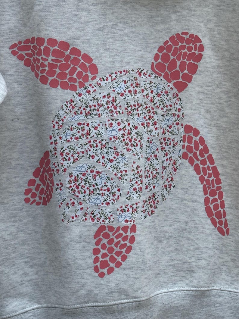 Puede incluir: Una sudadera gris con un dise&ntilde;o de tortuga marina. El caparaz&oacute;n de la tortuga tiene un estampado floral en rojo y blanco, mientras que las aletas y la cabeza son de color rojo liso. El dise&ntilde;o est&aacute; centrado en la parte delantera de la sudadera.