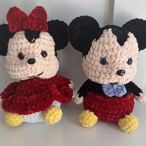 May include: Two plush crochet dolls, one Minnie Mouse and one Mickey Mouse. Minnie wears a red dress and bow, Mickey has red shorts and a blue bow tie. Both have black ears, black eyes, and yellow feet. The dolls are made of soft, textured yarn.